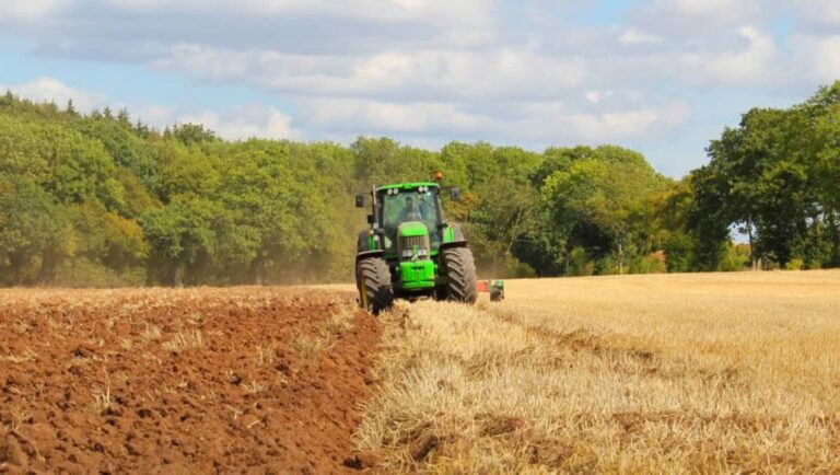 Green tractor ploughing field beside harvested crops